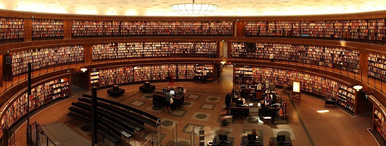 The circular reading room of a grand university library, with tiered wooden bookshelves and students seated at central desks.