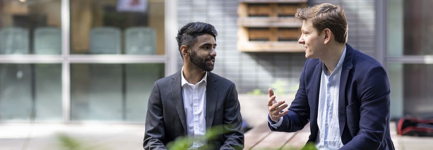 Two young professionals in business attire sitting outdoors and talking, one gesturing while speaking.