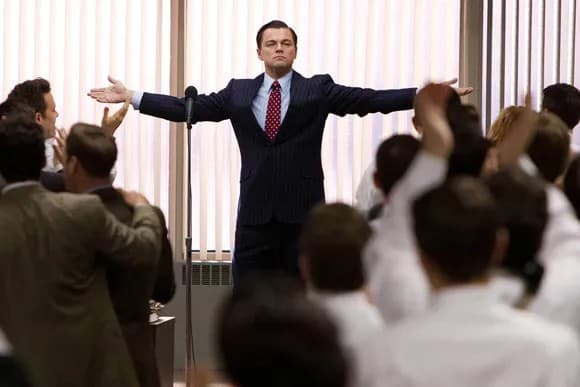 A man in a pinstriped suit standing at a podium with arms outstretched, addressing a large crowd of applauding colleagues in an office.