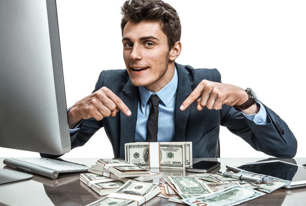A smiling young businessman in a suit pointing down at stacks of cash on his desk next to a computer monitor.