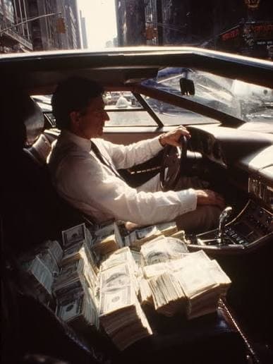 A man in a suit driving a convertible through a city street with stacks of cash piled on the passenger seat.