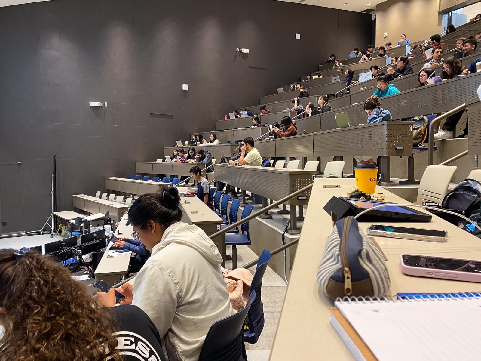 A packed, tiered college lecture hall full of students working on laptops and notebooks.