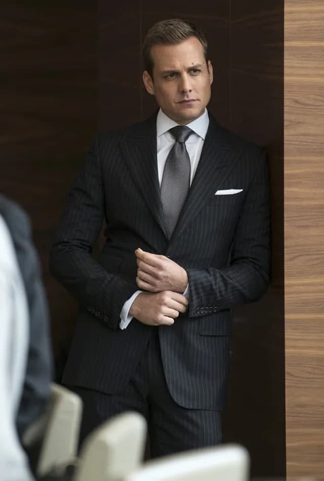 A well-dressed man in a pinstriped suit adjusting his cufflink while standing in a wood-paneled hallway.