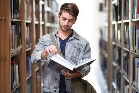 A college student standing between library shelves, reading an open book.