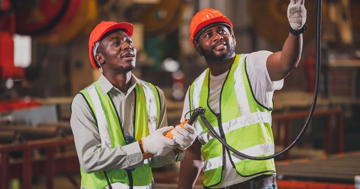 Two workers in hard hats and high-visibility vests training with industrial equipment on a factory floor, representing the hands-on workforce programs now eligible for Pell Grant funding.